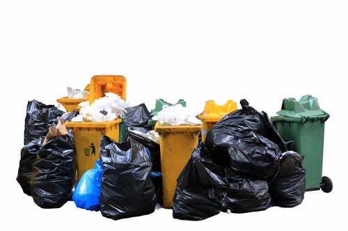A collection of various waste bins and black refuse bags arranged on a paved outdoor surface, with some bins being yellow, green, and orange, indicating different waste categories. The black bags appear to be filled with household or garden rubbish, stacked and leaning against the larger bins. The yellow bin and slightly visible orange bin are closed, while the green bin is slightly open, revealing some waste inside. The scene is set outdoors in natural daylight, likely in a residential area, with no background scenery visible. The arrangement suggests a waste collection or rubbish disposal situation, relevant to services offered by Garden Clearance Hampstead for rubbish removal.