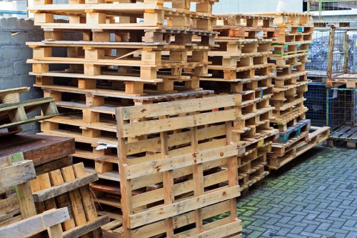 A large stack of wooden pallets is arranged outdoors on a paved surface, with some pallets leaning against a brick wall, and others stacked neatly in multiple layers. The pallets are light brown with visible wood textures and vary slightly in size and condition, some showing minor wear or discoloration. To the left, there is a smaller wooden structure, possibly a partially assembled pallet or a piece of outdoor furniture, resting on the ground. In the background, there is a chain-link fence and some metal scaffolding, indicating an industrial or construction area, potentially near Hampstead. The scene is well-lit with natural daylight, highlighting the textures of the wood and the surrounding environment. The image depicts materials that are typical of waste or rubbish collection services, which Garden Clearance Hampstead might manage as part of their rubbish removal offerings in the local area.