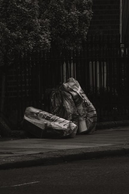 A person standing outdoors on a grassy surface holding a large black garbage bag open, preparing for rubbish collection. They are wearing a green long-sleeve shirt, a yellow and gray checkered flannel